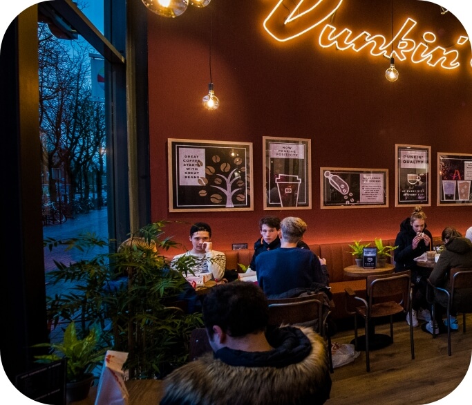 People are seated at tables inside a brightly lit café with a neon "Dunkin'" sign on the wall. The atmosphere is cozy, reminiscent of a coffee and donuts franchise, with vibrant indoor plants and framed artwork decorating the space.