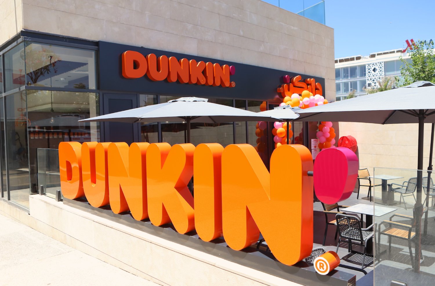 Outdoor seating area in front of a Dunkin' coffee and donuts franchise, featuring large orange Dunkin' letters, umbrellas, and colorful balloons.