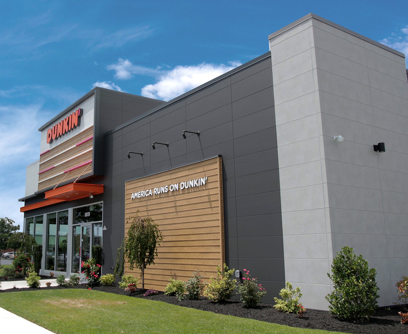 A modern Dunkin' building with a gray and beige exterior and a sign reading "America Runs on Dunkin'." As a coffee and donuts franchise, the storefront features large windows and landscaped greenery.