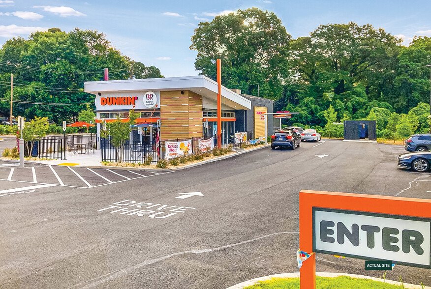         A Dunkin' coffee and donuts franchise with a drive-thru lane and a parking lot, surrounded by greenery and trees. An "ENTER" sign is visible in the foreground. Several cars are lined up at the drive-thru.