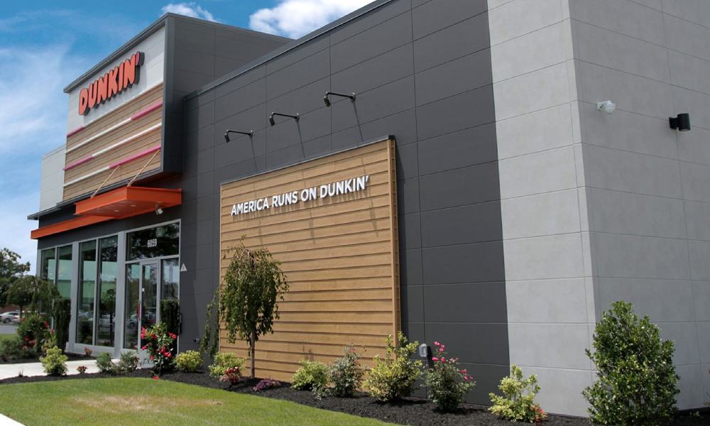 The exterior of this Dunkin' store, part of the popular coffee and donuts franchise, features the "America Runs on Dunkin'" slogan on the wall, black and grey panels, and a glass entrance. It is surrounded by landscaped bushes and plants under a blue sky.