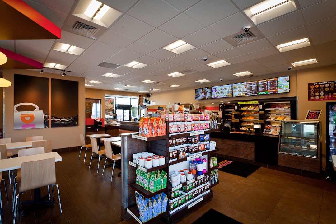 Interior of a coffee and donuts franchise featuring shelves of beverages and snacks, a counter area displaying menu boards, baked goods, and welcoming seating arrangements with tables and chairs.